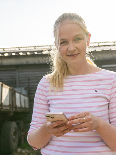 Landwirtin Vanessa Schneider hält ihr Smartphone in der Hand und lächelt.